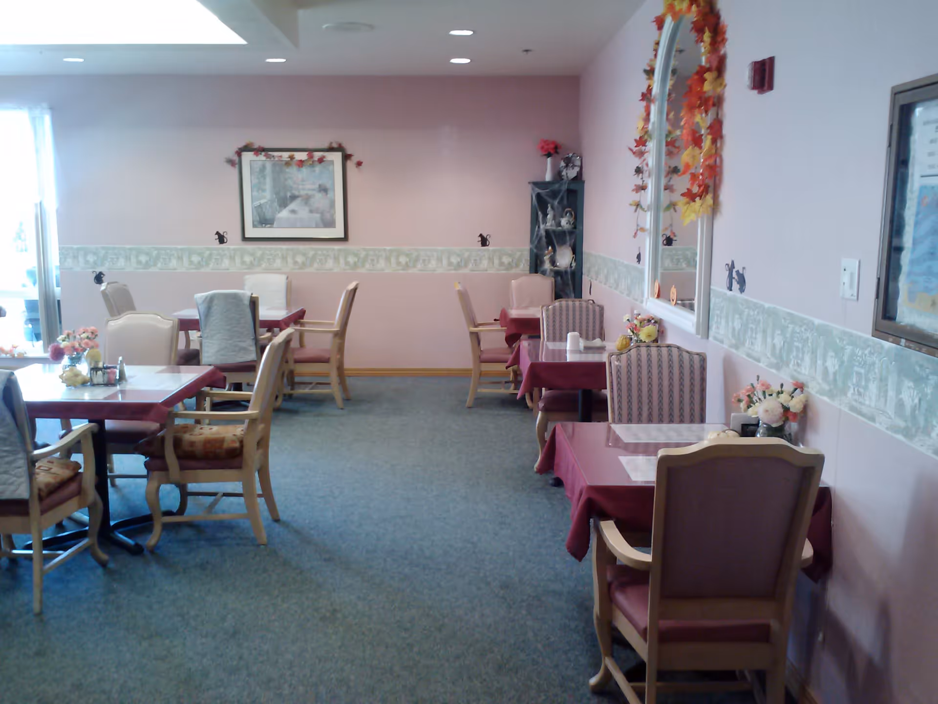 A dining room with several tables covered with maroon tablecloths and surrounded by wooden chairs with cushions. The walls are painted light pink with a decorative green and white border. There is a large mirror on the right wall adorned with autumn leaf decorations, a framed picture on the far wall, and a small shelving unit with decorative items in the corner. The room is carpeted and well-lit with natural light coming from a window on the left.