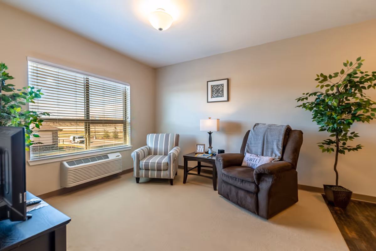 A cozy living room with a large window with blinds, a striped armchair, a brown recliner with a 'Home Sweet Home' pillow, a small side table with a lamp and decorative items, a potted plant, and a TV on a stand.