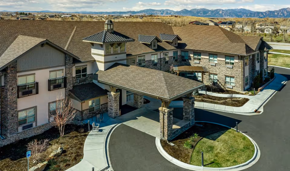Aerial view of a two-story senior living building with a covered porte-cochere entrance, stone and siding exterior, and surrounding drive and landscaping.