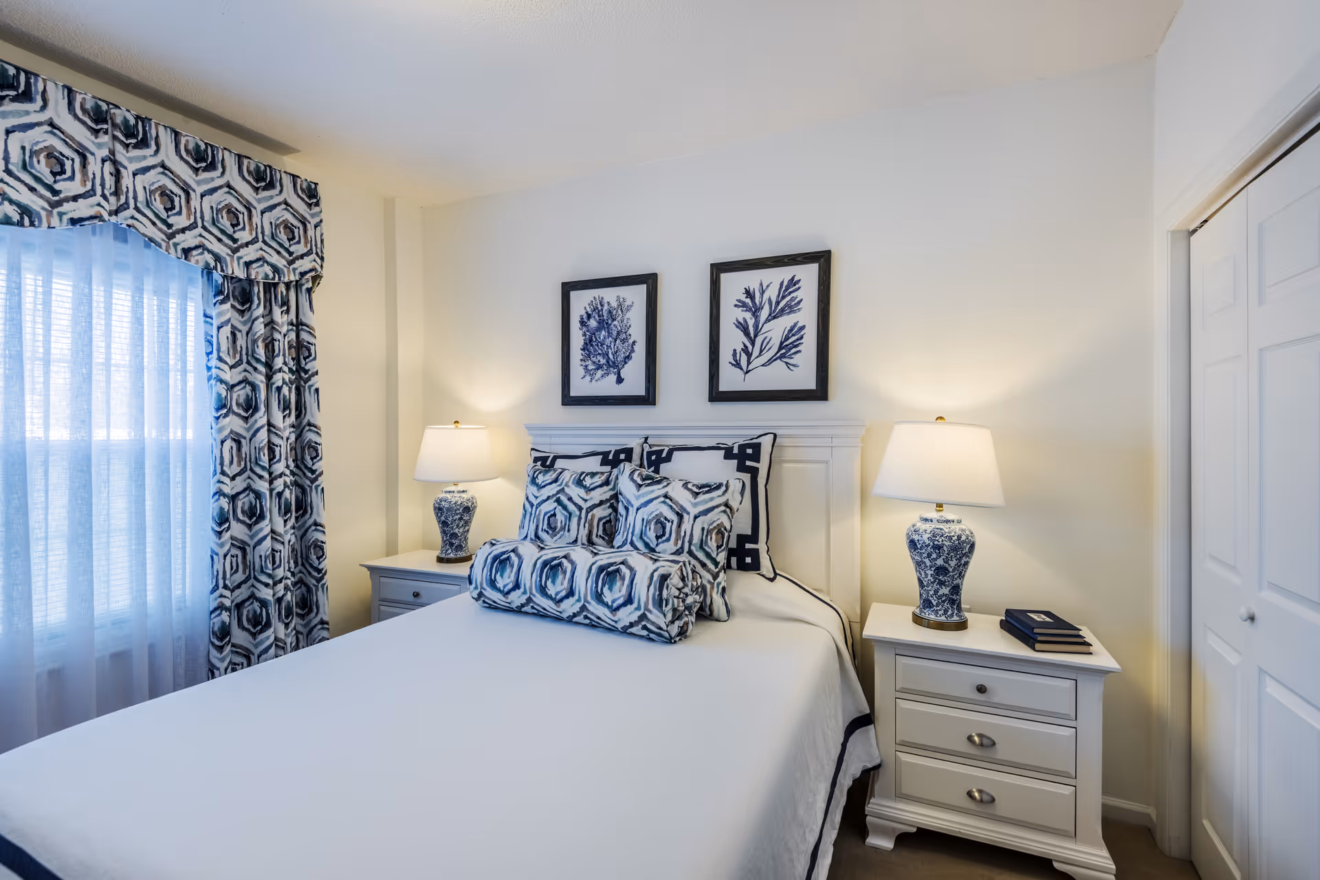 A neatly made bed with white bedding and blue patterned pillows and bolster in a bedroom. Two matching bedside tables with blue and white lamps flank the bed. Above the bed are two framed botanical prints. A window with blue and white patterned curtains is on the left, and a white closet door is on the right.