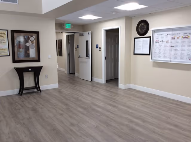 Interior hallway of a senior living facility with light wood flooring, beige walls, a small black table with a framed artwork above it on the left wall, an open door leading to another room, a clock and framed documents on the right wall, and an exit sign above a door in the background.