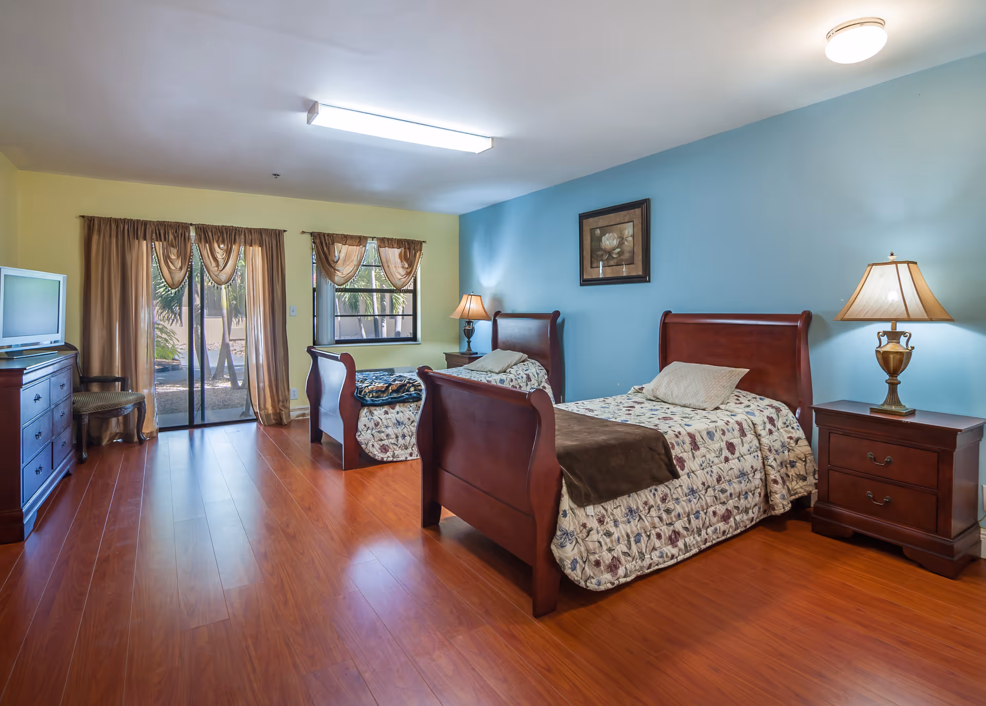 A senior living bedroom with two wooden twin beds, each with floral patterned bedspreads and pillows. The room has wooden flooring, a blue accent wall behind the beds, and a yellow wall with a window and sliding glass door covered by brown curtains. There are two bedside tables with lamps, a wooden dresser with a TV on top, and a chair next to the dresser.
