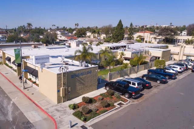 Aerial view of Shoreline Healthcare Center, a single-story building with a flat white roof, surrounded by palm trees and a parking lot with several cars. The building is located in a suburban area with houses and trees in the background.
