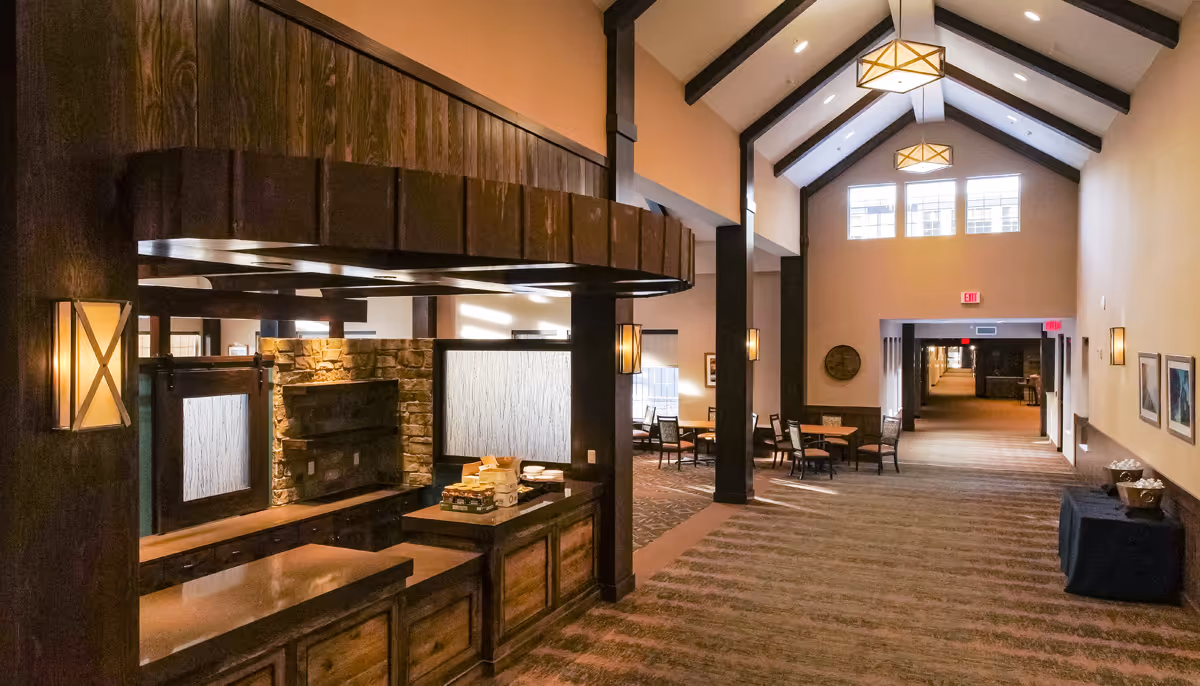 Spacious senior living facility interior with a vaulted ceiling, wooden reception/serving counter, seating area, and a long carpeted hallway.