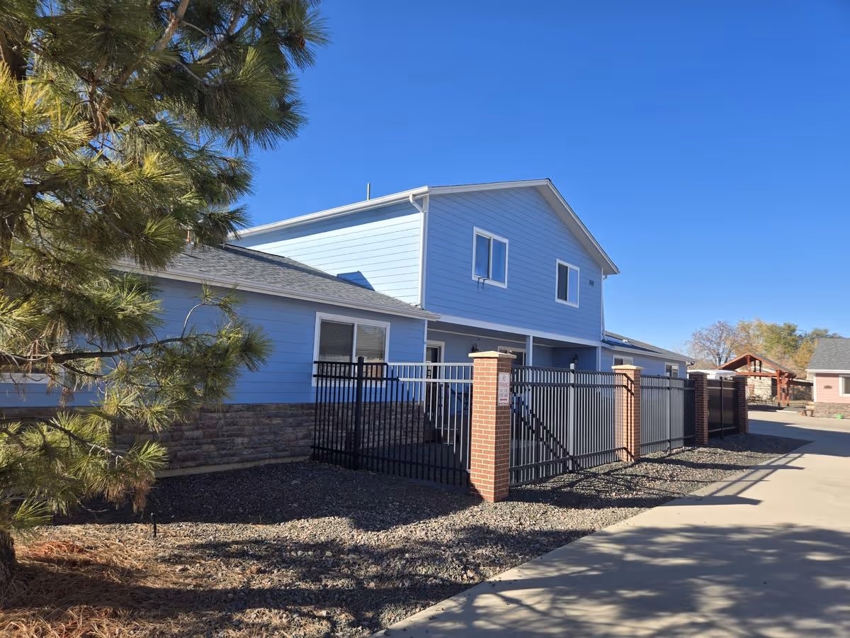 Exterior view of a two-story light blue building with a fenced area, driveway, and a tree under a clear blue sky.