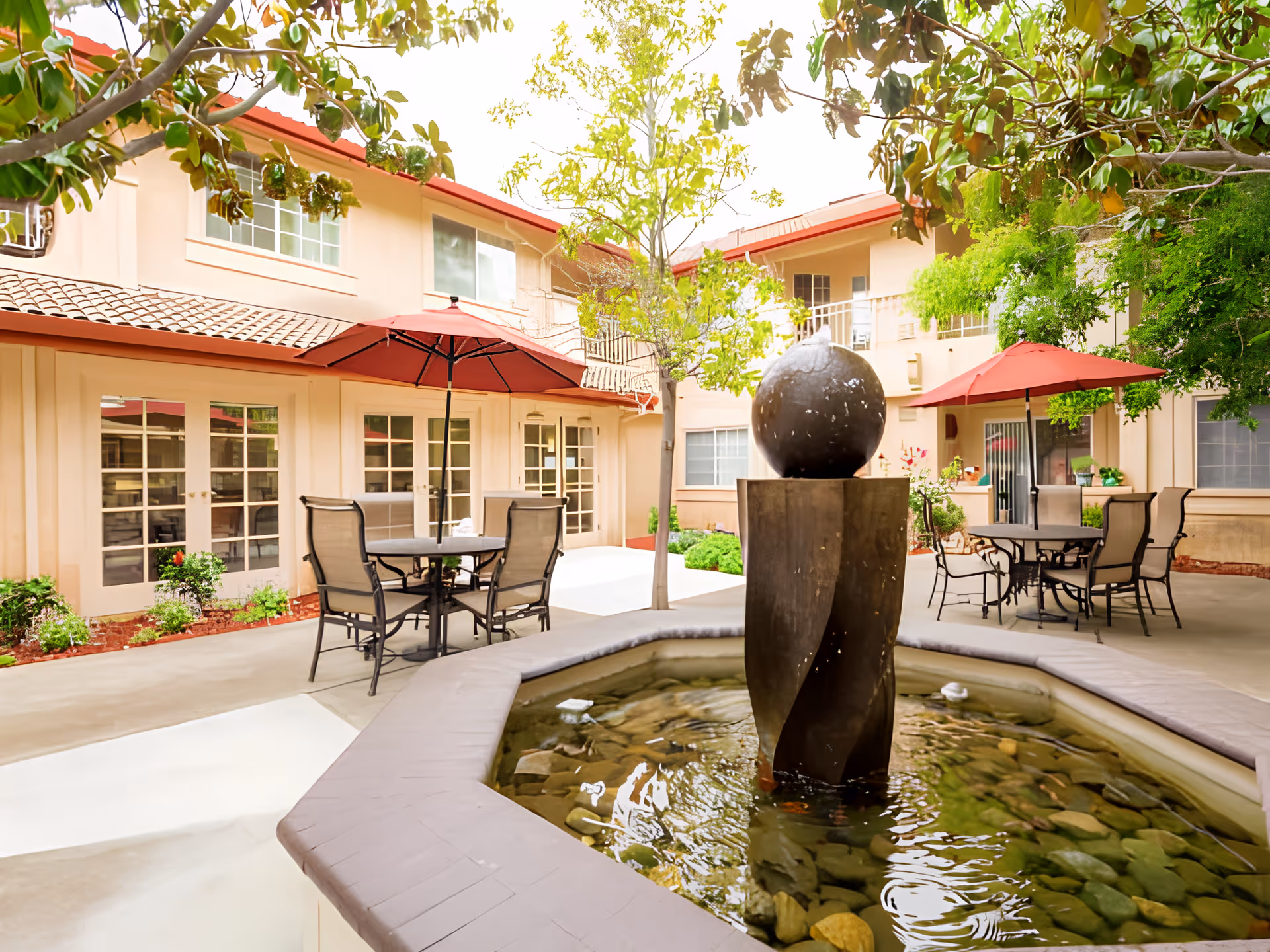 Outdoor courtyard area of a senior living facility with a central water fountain surrounded by a stone basin. There are patio tables with chairs and red umbrellas, trees, and a two-story building with beige walls and red roof tiles in the background.