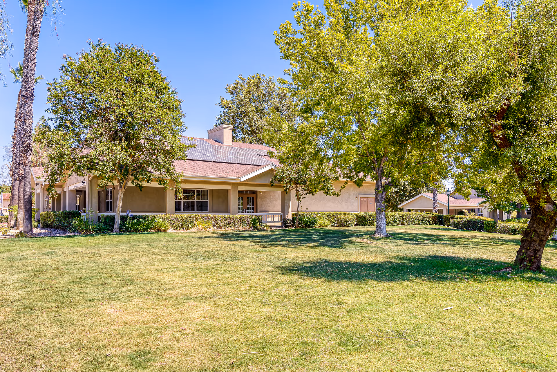 Exterior view of a single-story building with a sloped roof and solar panels, surrounded by green trees and a well-maintained grassy lawn under a clear blue sky.