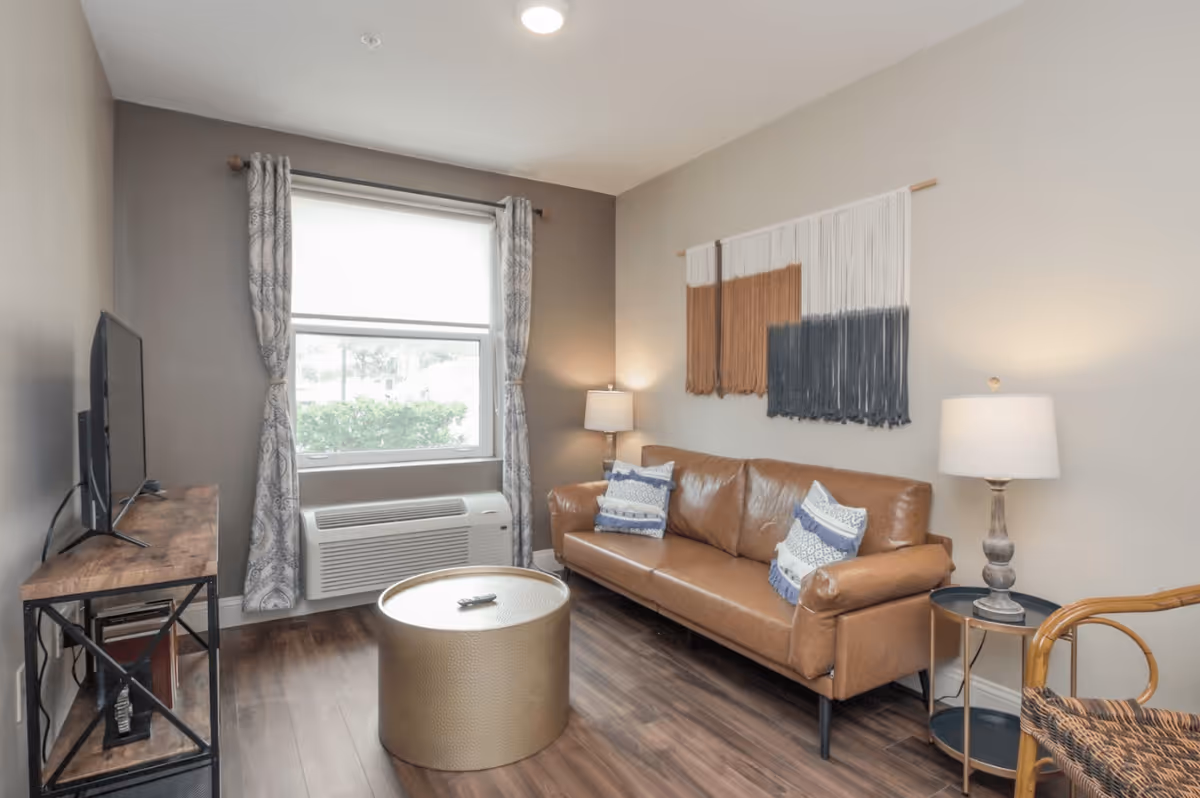 Living room with a brown leather sofa, round gold coffee table, TV on a console, window with curtains, and wall hanging.