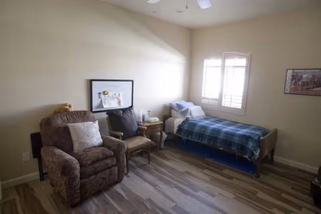 A small, cozy room with a single bed covered with a blue plaid blanket near a window with white shutters. Next to the bed is a wooden nightstand with a lamp and some items on it. There is a cushioned armchair with a white pillow and a wooden chair with a dark cushion. The walls are light-colored, and the floor has wood-patterned vinyl or laminate flooring. A framed picture and a whiteboard are hung on the walls.