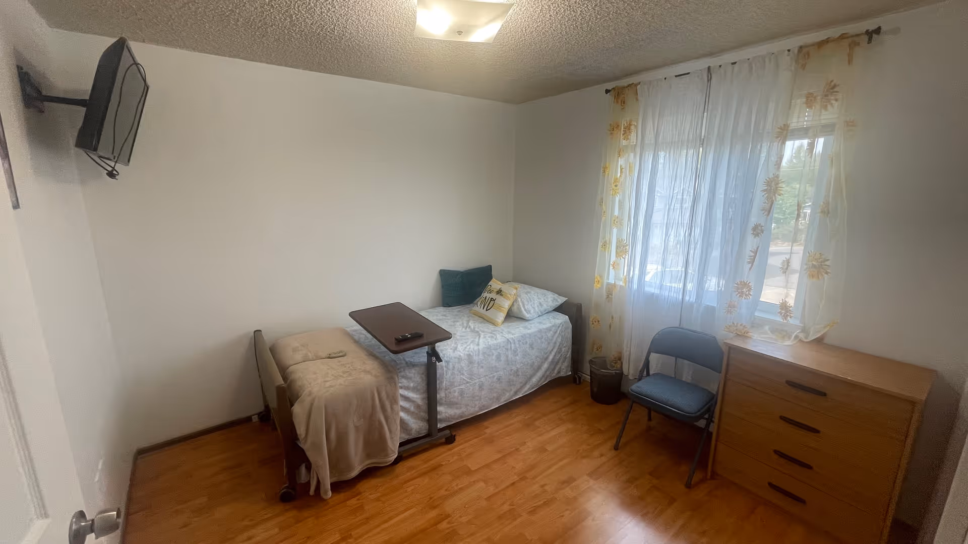 A small bedroom with a single bed covered with a light-colored bedspread and pillows, including one decorative pillow. There is a beige blanket draped over the foot of the bed and a small overbed table positioned on top. A flat-screen TV is mounted on the wall opposite the bed. The room has wooden flooring, a window with sheer white curtains featuring a yellow floral pattern, a blue folding chair, and a wooden dresser with three drawers.