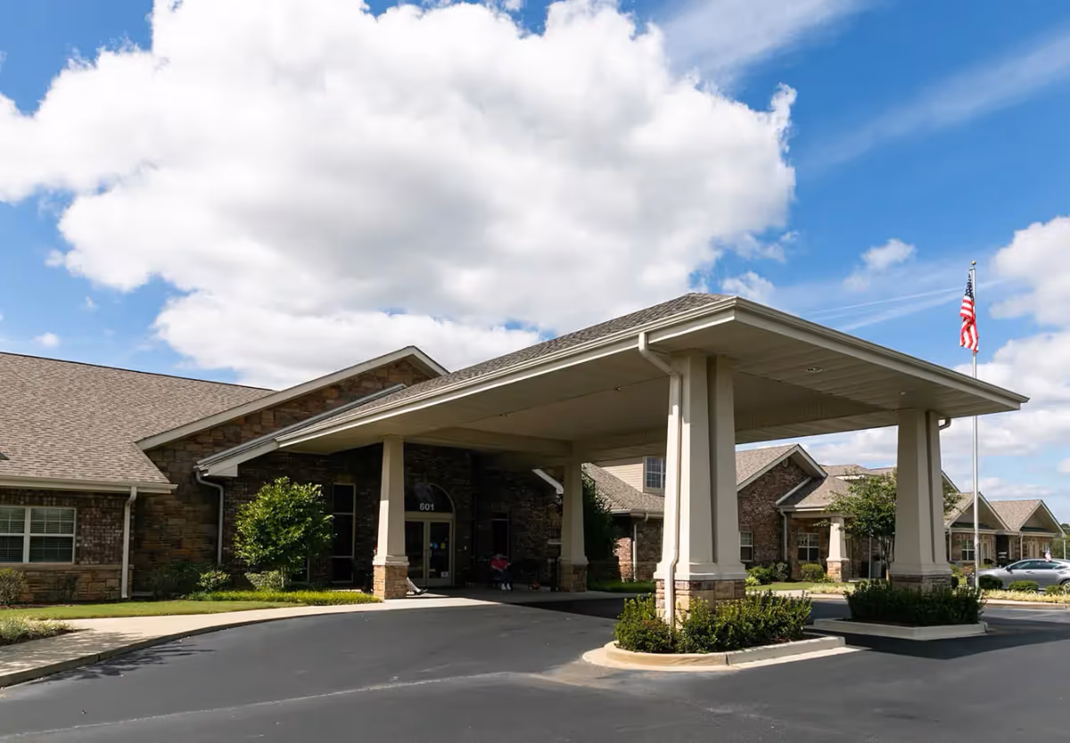 Exterior view of Hearthside Senior Living facility showing the main entrance with a covered drop-off area supported by columns, brick and stone facade, a driveway, landscaped bushes, and an American flag on a flagpole under a partly cloudy blue sky.