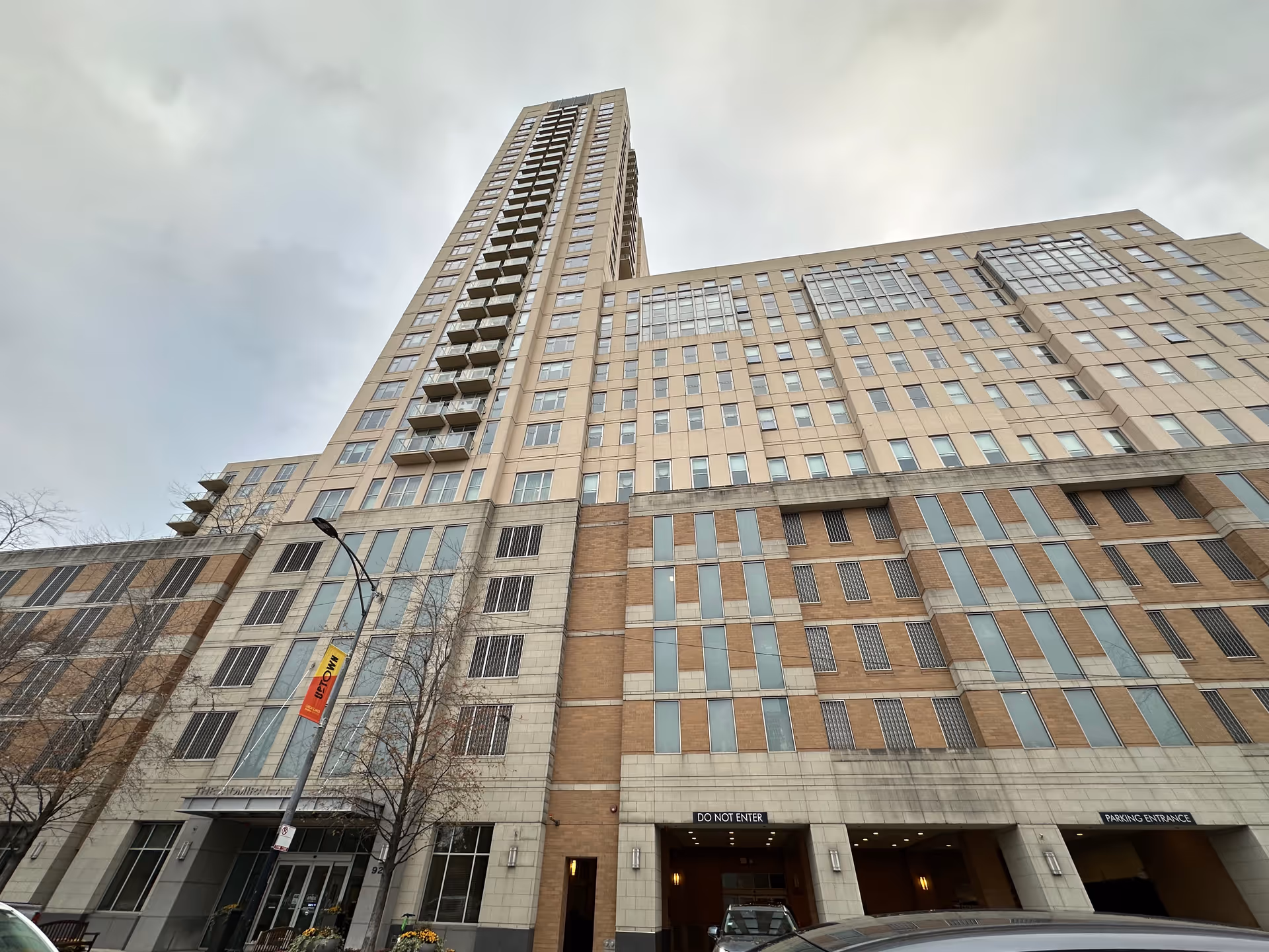 Low-angle view of a tall beige high-rise residential building with balconies, street-level entrance and parking garage.