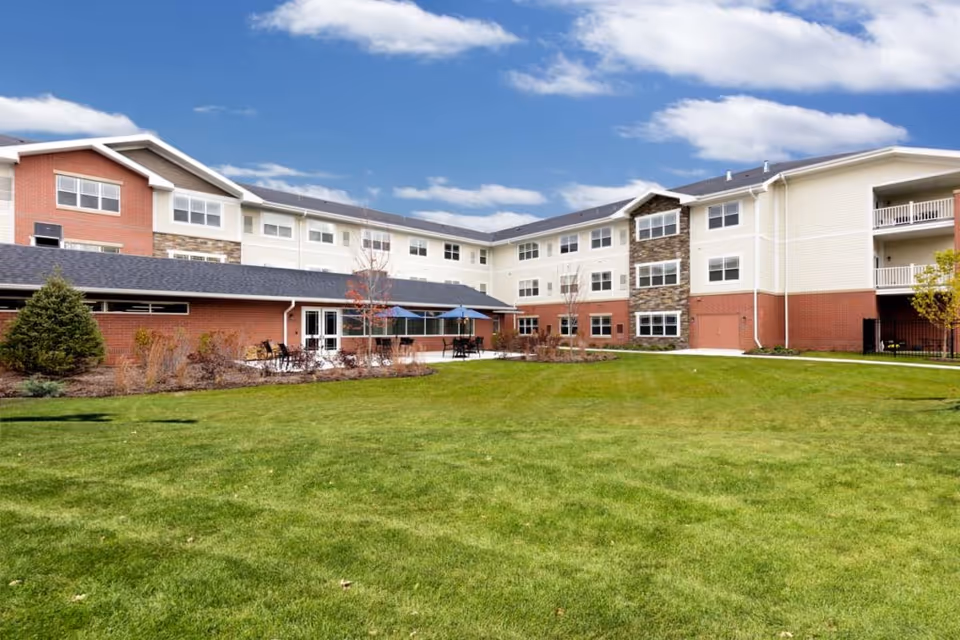 Three-story red-and-white senior living building with a large green lawn and outdoor patio seating under a blue sky.
