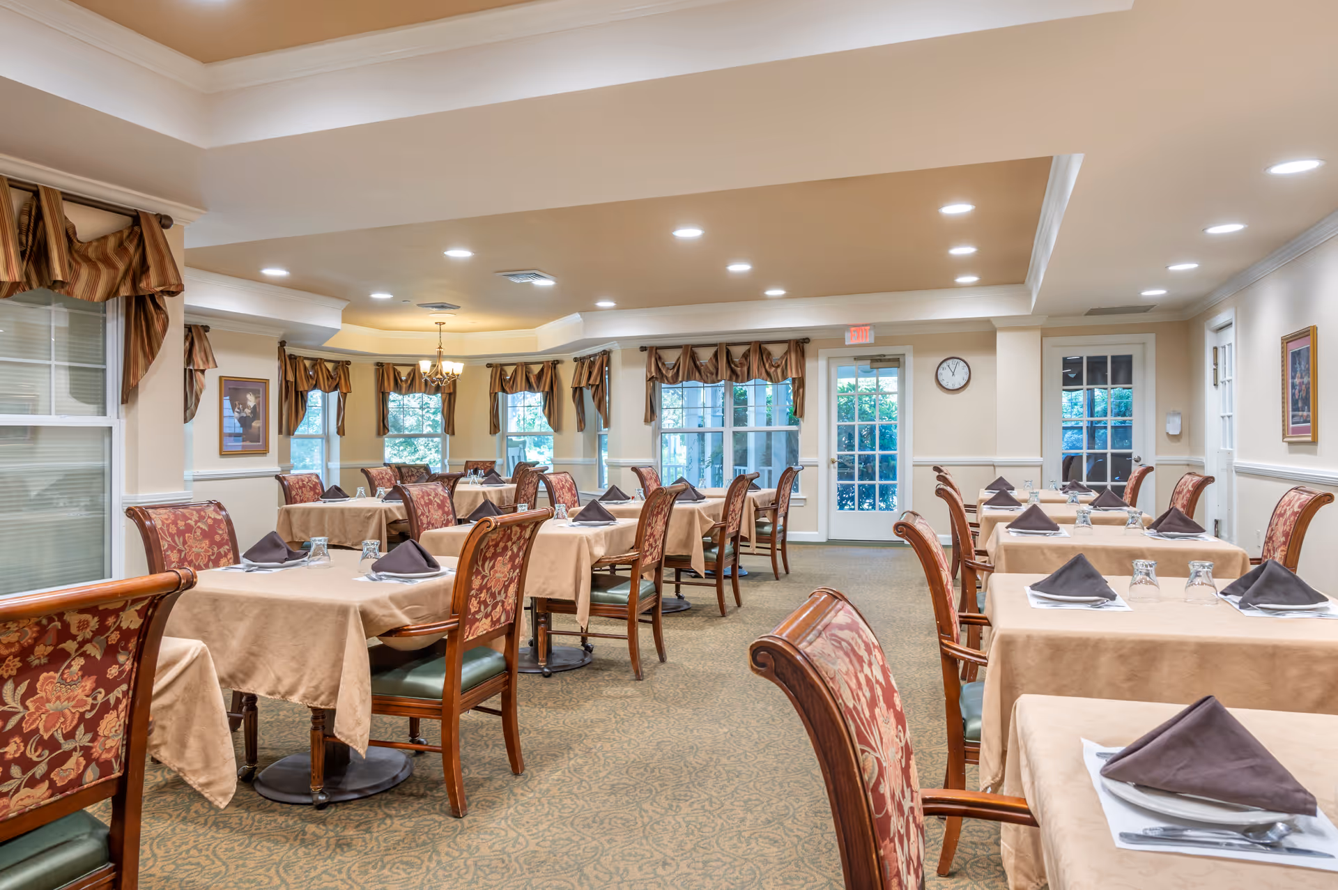 A well-lit dining room with multiple tables covered in beige tablecloths, each set with plates, silverware, glasses, and folded brown napkins. The chairs have wooden frames with floral upholstery. Windows with brown valances line the walls, and there is a door with glass panes at the far end. The ceiling has recessed lighting and a chandelier.