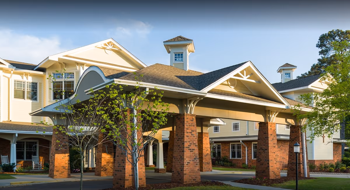 Covered entrance with brick pillars and a porte-cochère at the front of a multi-story senior living building with surrounding trees.