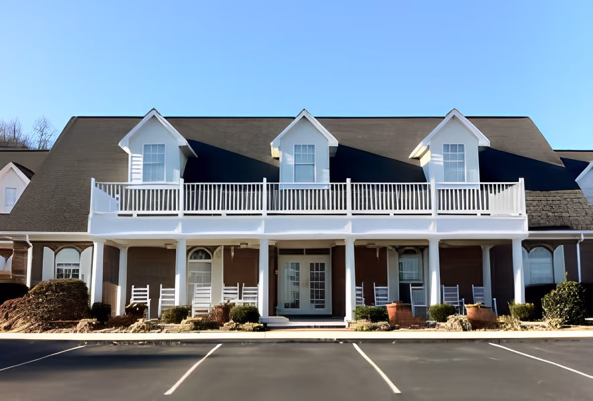 Front exterior view of a senior living facility building with a large porch featuring white railings and several white rocking chairs. The building has a dark roof with three dormer windows and a clear blue sky in the background. There is a parking lot in front of the building.