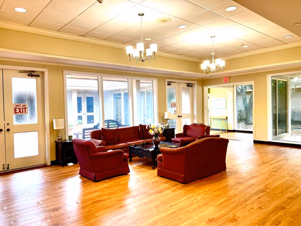 A spacious and well-lit common area with wooden flooring, featuring a seating arrangement of red upholstered sofas and armchairs around a dark wooden coffee table with a flower vase. The room has large windows and glass doors providing natural light and views of an outdoor courtyard. Two chandeliers hang from the ceiling, and there are side tables with lamps near the doors.