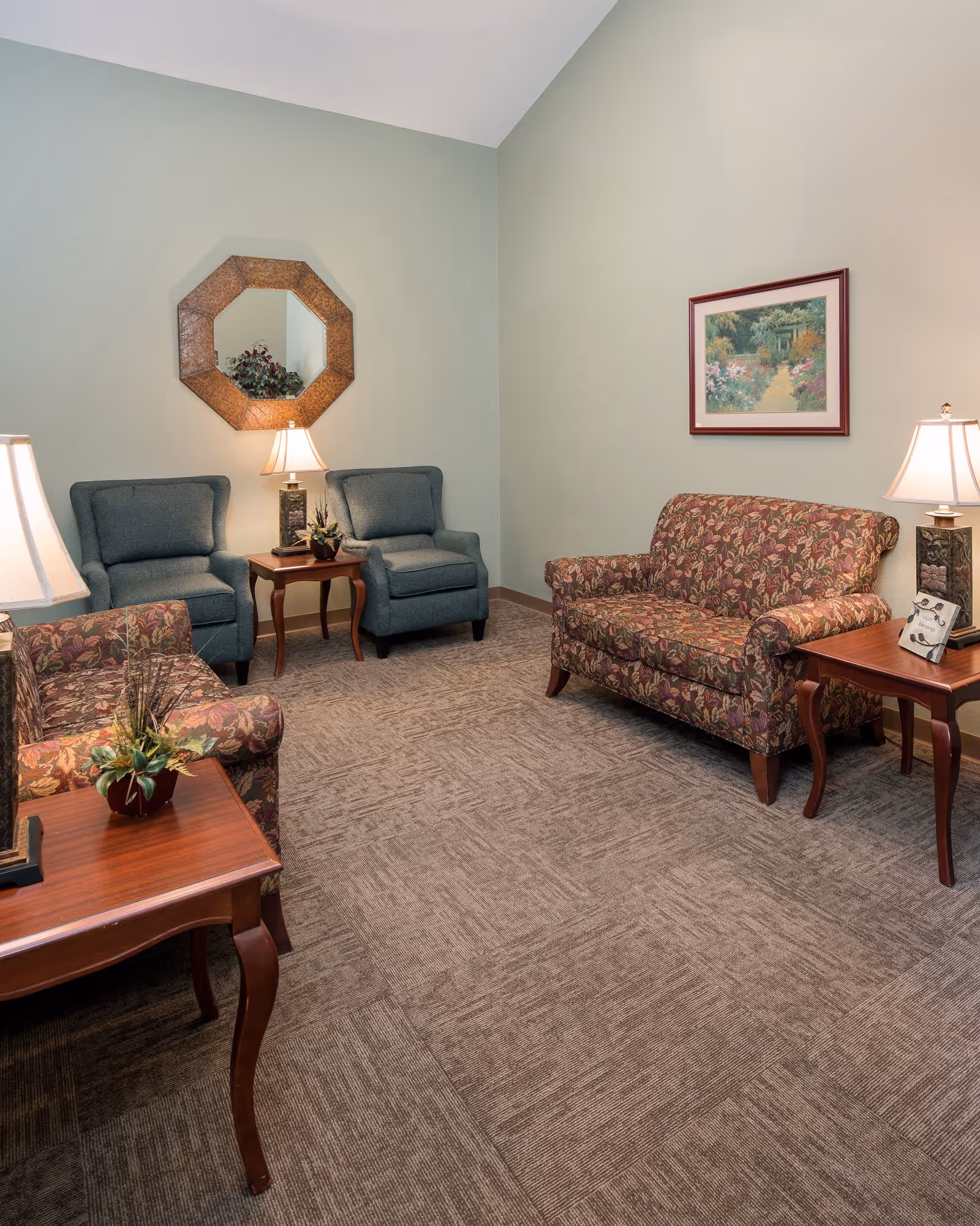 A cozy sitting room with patterned sofas, two blue armchairs, side tables and lamps against a pale green wall with a mirror and framed painting.