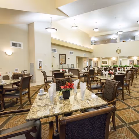 A spacious dining room with multiple tables set with white napkins, cups, and silverware. Each table has a small red flower centerpiece. The room features high ceilings, warm lighting, and a patterned carpet floor. There are chairs with dark wood frames and upholstered seats around the tables. The walls are decorated with framed pictures and a clock is visible on the far wall.