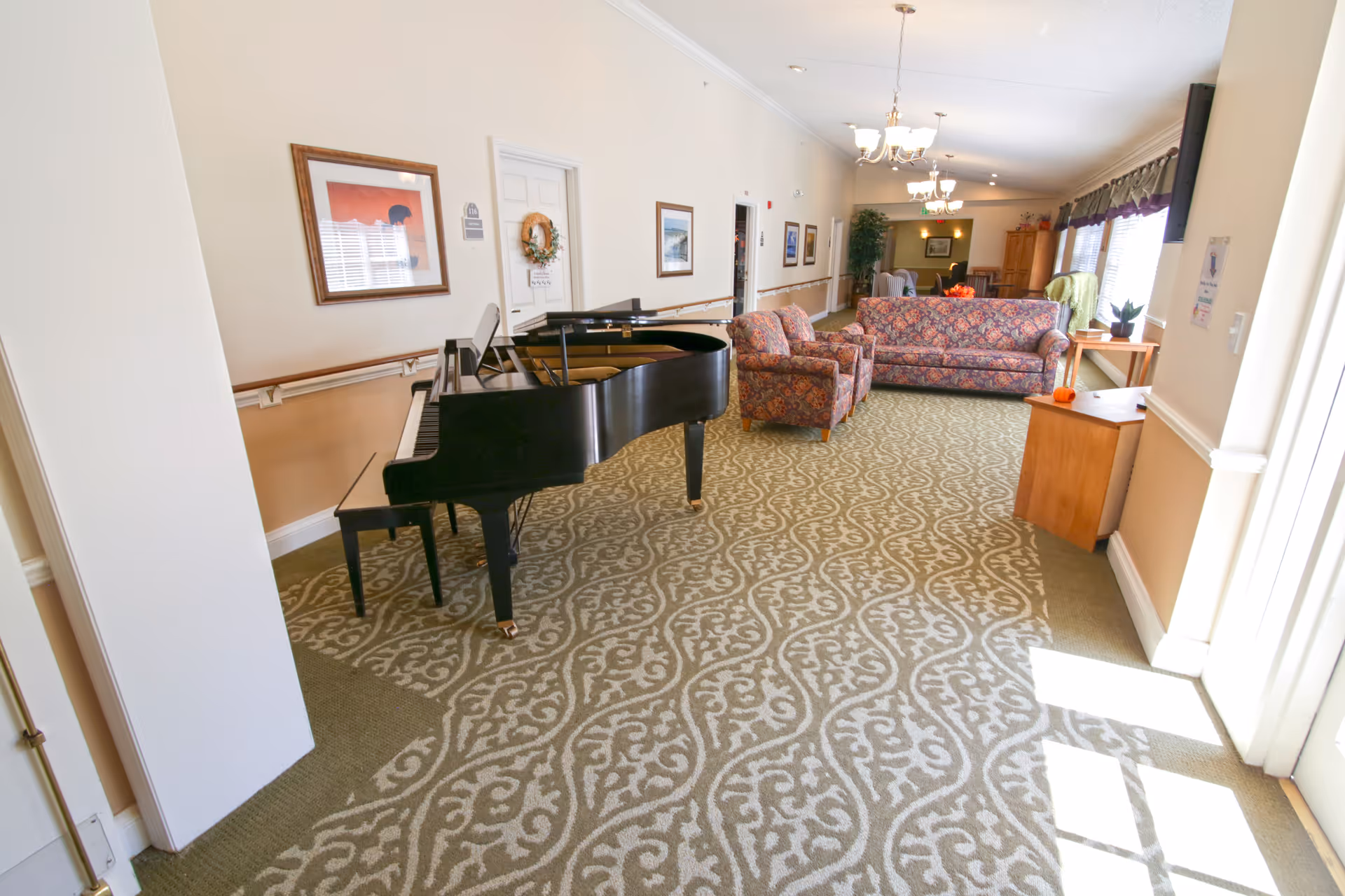 A bright and spacious common area in a senior living facility featuring a black grand piano with a bench, a patterned armchair, and a matching floral sofa. The room has patterned carpet flooring, cream-colored walls adorned with framed artwork, and several ceiling light fixtures. Large windows on the right side allow natural light to fill the space, and there is a small wooden table with a plant and a pumpkin decoration near the windows.