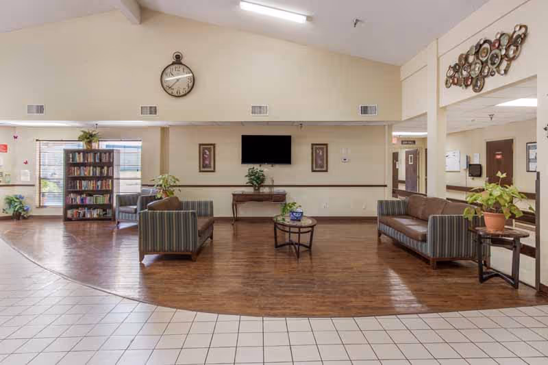 A spacious common area in a senior living facility with wooden flooring and tiled entrance. The room features two striped armchairs and a brown leather sofa arranged around a round glass coffee table with a plant on it. There is a bookshelf filled with books near a window, a wall clock above a mounted flat-screen TV, and decorative framed pictures on the wall. Several potted plants and wall decorations add a homey touch.