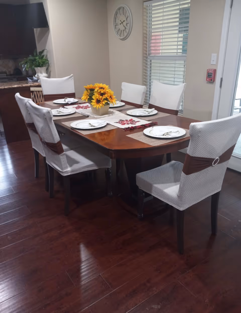 A dining room with a wooden table set for six, white-covered chairs and a sunflower centerpiece on dark hardwood floors.