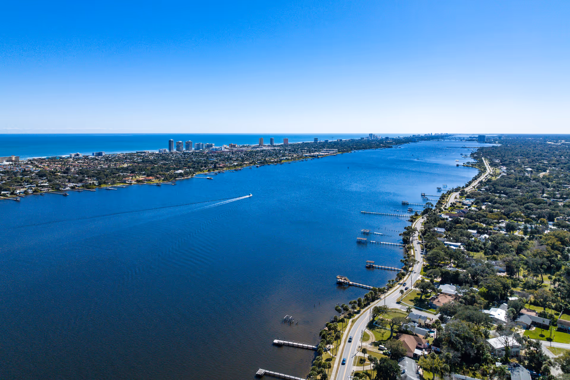 Aerial view of a coastal city with a large body of water on the left and a road running alongside it on the right. The shoreline features docks extending into the water, residential houses, and greenery. In the distance, there are tall buildings near the horizon under a clear blue sky.