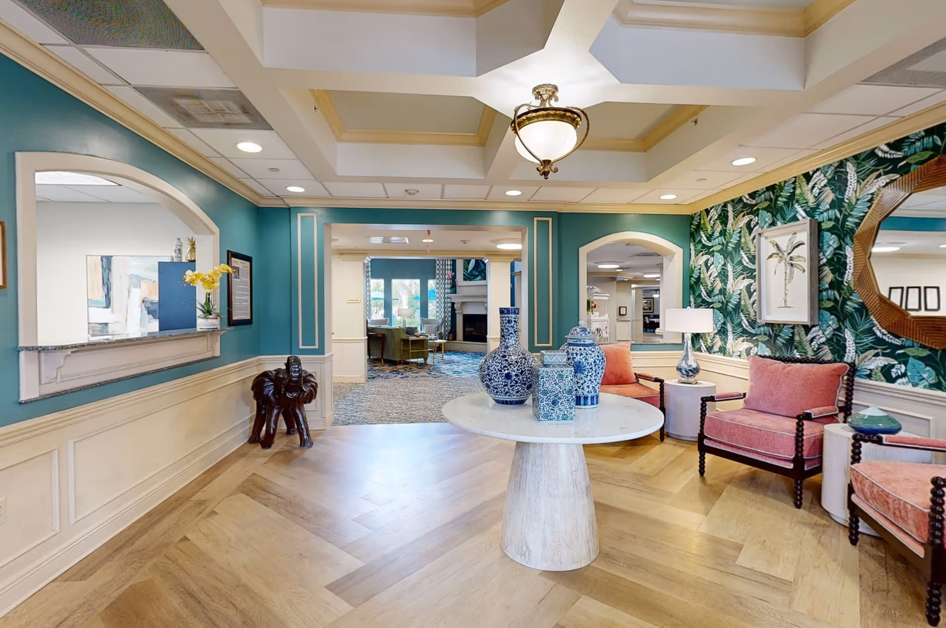A stylish and welcoming interior common area with teal walls, a coffered ceiling with a central light fixture, and a round white table displaying blue and white decorative vases. The room features pink upholstered chairs, a side table with a lamp, tropical leaf wallpaper, framed artwork, and a wooden elephant sculpture near a pass-through window.