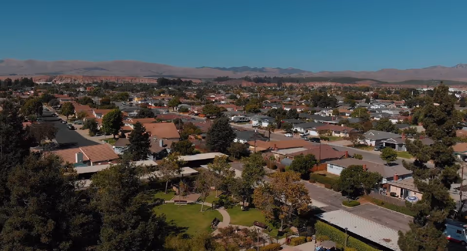 Aerial view of a suburban neighborhood with numerous houses, trees, and streets under a clear blue sky with mountains in the background.