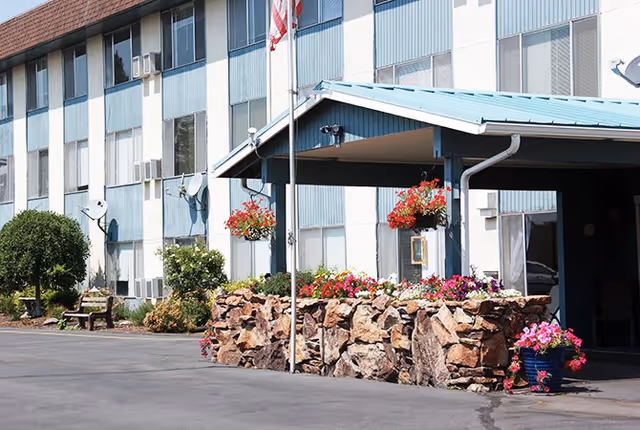 Front entrance of a multi-story senior living building with a covered drop-off, rock planters and hanging flower baskets.