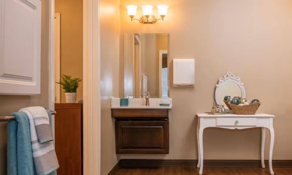 Interior view of a bathroom area with a wall-mounted sink and mirror under a three-light fixture. To the right, there is a white vanity table with an ornate mirror and a basket filled with decorative balls. A towel rack with folded towels is visible on the left side.