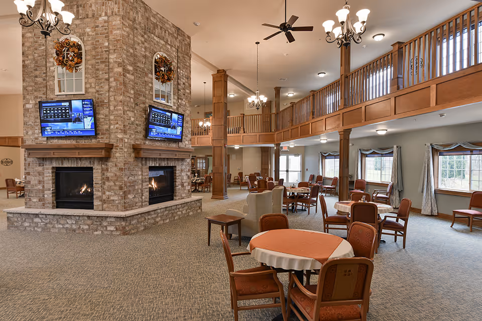 Spacious common area in a retirement community featuring a large brick double-sided fireplace with two mounted flat-screen TVs above it. The room has multiple round tables with chairs, comfortable armchairs, carpeted floor, and large windows with curtains allowing natural light. There is a wooden balcony railing on the upper level and several ceiling light fixtures.