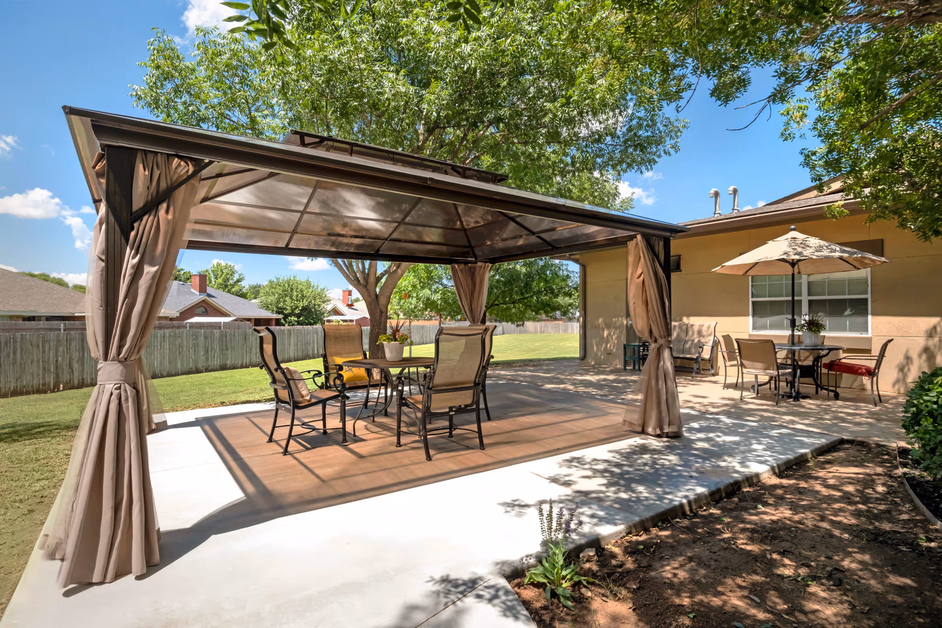 Outdoor patio area with a metal gazebo covering a table and four chairs. Nearby, there is another table with an umbrella and additional chairs. The area is surrounded by a wooden fence, trees, and a grassy lawn under a clear blue sky.