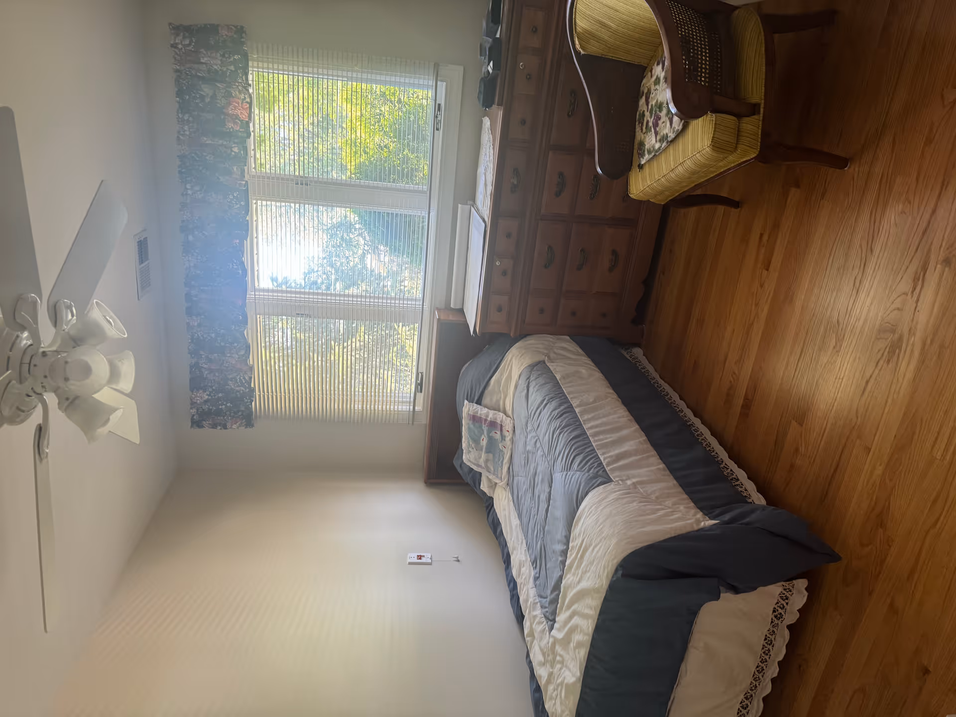 Bedroom with bed, dresser, chair, and window blinds in a senior living facility.