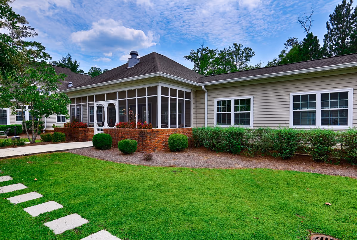Exterior view of a single-story building with beige siding and white-framed windows, featuring a screened-in porch with a white door. The building is surrounded by neatly trimmed bushes, a green lawn, and a stone pathway leading to the entrance. Trees and a partly cloudy sky are visible in the background.