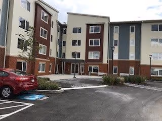 Exterior view of a multi-story senior living facility building with a parking lot in front, including a red car parked in a handicapped parking space. The building has a combination of beige, brown, and gray colors with multiple windows and entrances.