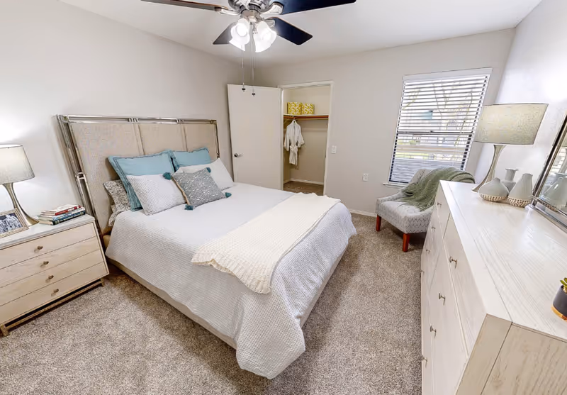 Bright furnished bedroom with a bed, nightstand and lamp, dresser, chair, and an open closet.