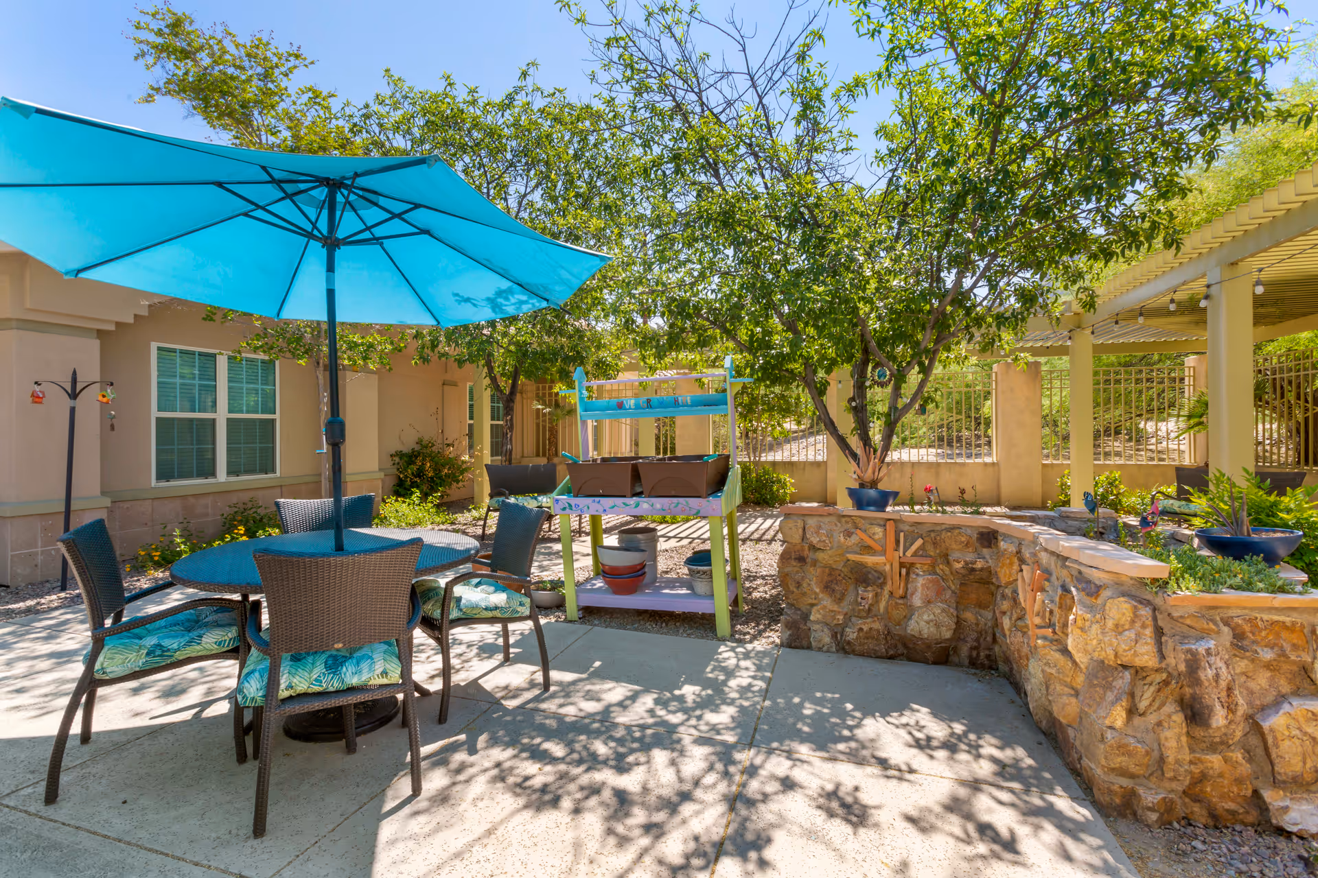 Outdoor patio area with a round table and four chairs with blue patterned cushions under a large blue umbrella. There are trees, a stone planter, and a small wooden shelf with potted plants. The area is sunny with shadows cast on the concrete ground.