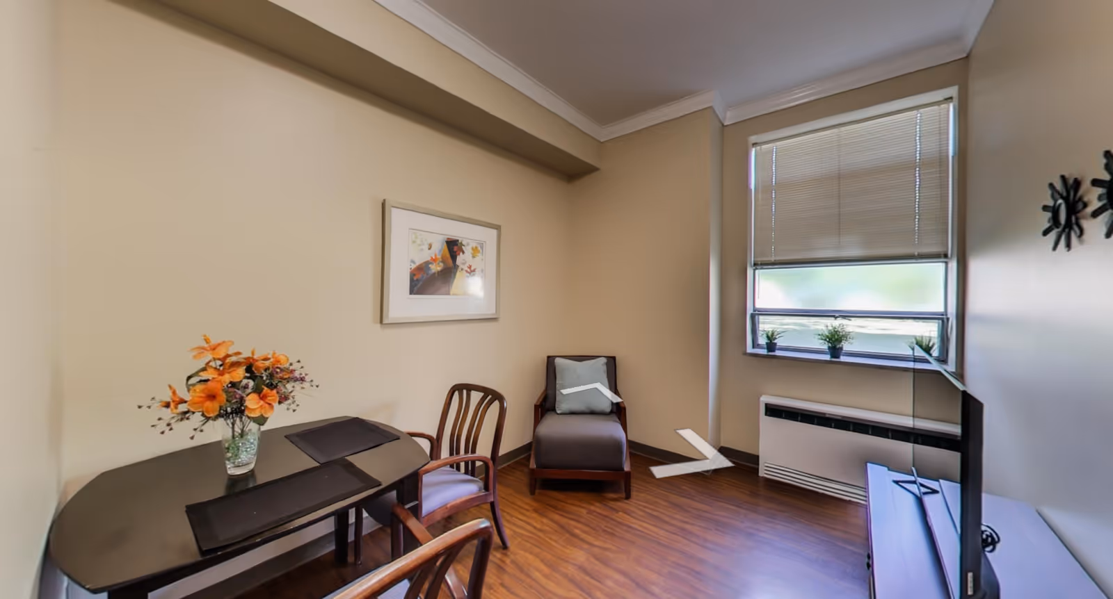 A small room with beige walls and wood flooring featuring a dark wooden table with two chairs, a vase with orange flowers on the table, a cushioned armchair with a pillow, a window with blinds partially closed and three small potted plants on the windowsill, a flat-screen TV on a stand, and a framed picture on the wall.