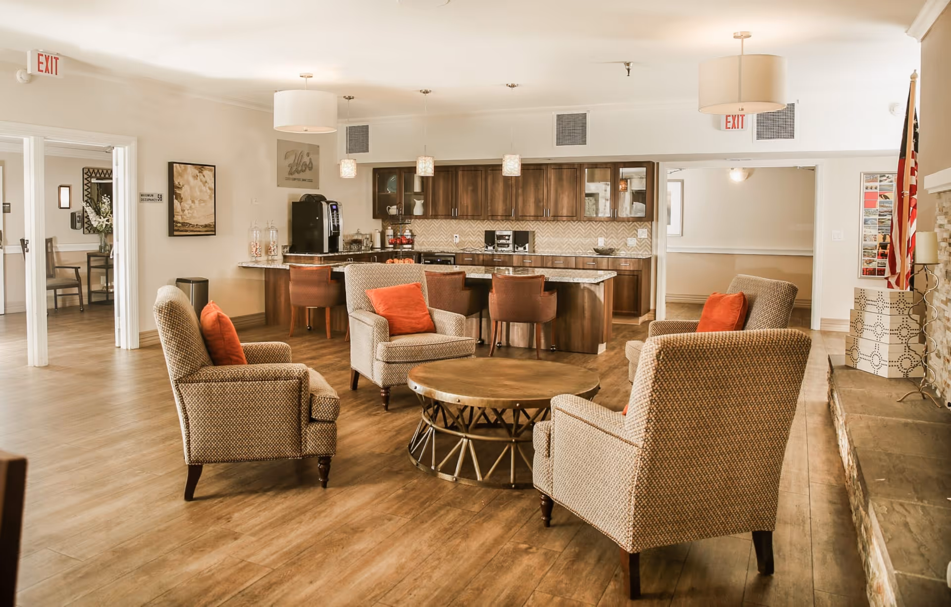 A cozy senior living common area with four patterned armchairs each with an orange cushion arranged around a round wooden coffee table. Behind the seating area is a kitchen with dark wood cabinets, a granite countertop island with four brown chairs, and various kitchen appliances. The room has wooden flooring, pendant lights, and exit signs above doorways. An American flag stands near a stone fireplace on the right.