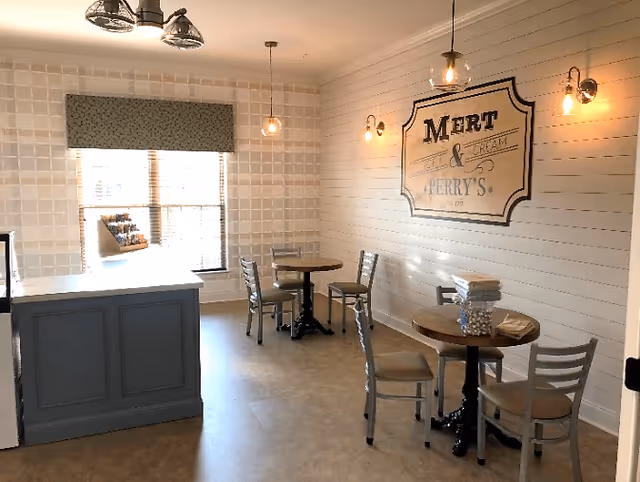 A small dining area with two round tables, each surrounded by three chairs. The walls are decorated with white shiplap and a large vintage-style sign that reads 'Mert & Perry's'. There are two hanging pendant lights and two wall sconces providing warm lighting. A window with a patterned valance lets in natural light, and a counter or service area is visible on the left side of the room.
