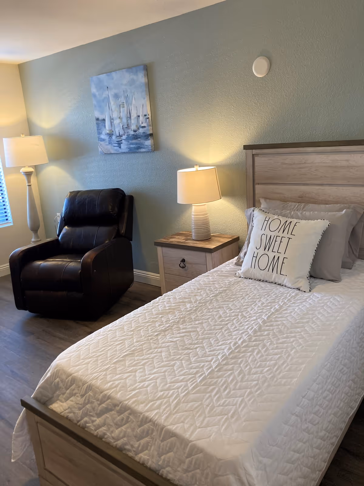 A tidy bedroom with a single bed topped by a 'Home Sweet Home' pillow, a nightstand and lamp, and a leather recliner against a light green wall.