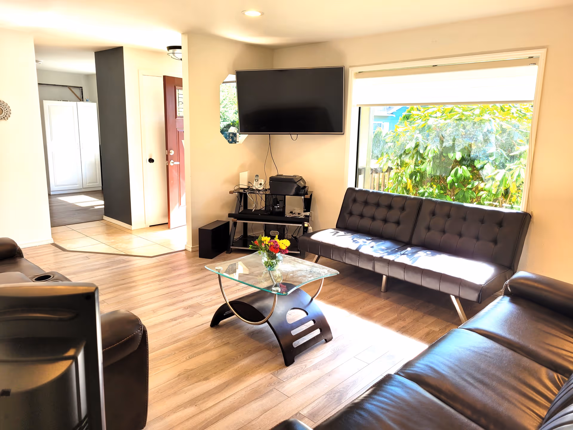 A bright living room with wooden flooring, featuring a black leather sofa and a black tufted couch. A glass coffee table with a vase of flowers sits in the center. A flat-screen TV is mounted on the wall above a small black entertainment unit with electronic devices. A large window lets in natural light and shows green foliage outside.