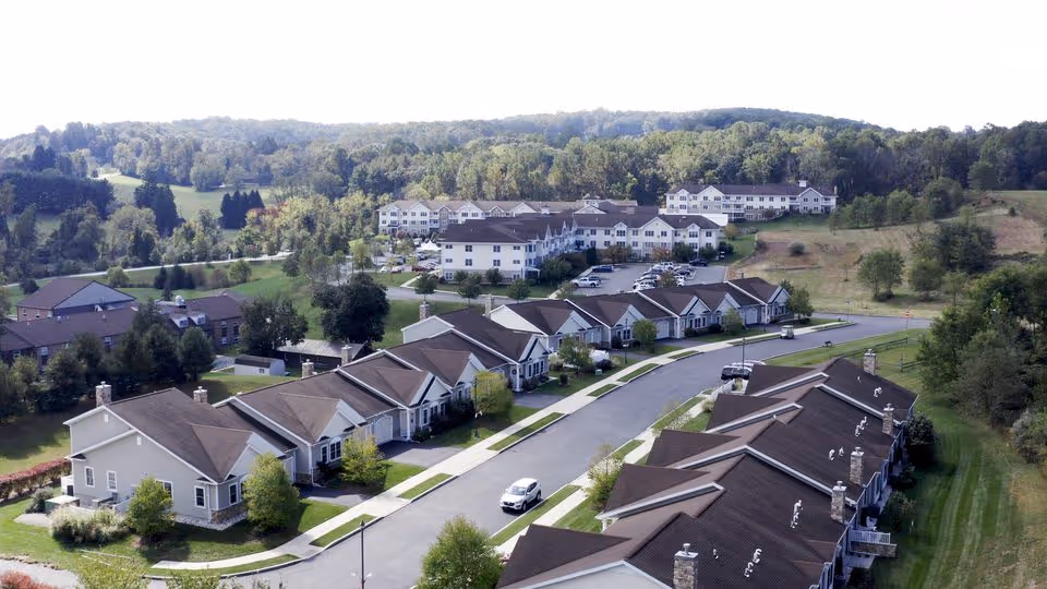 Aerial view of a senior living community with rows of single-level homes, a curving road, parking areas, and larger buildings set among trees and rolling hills.