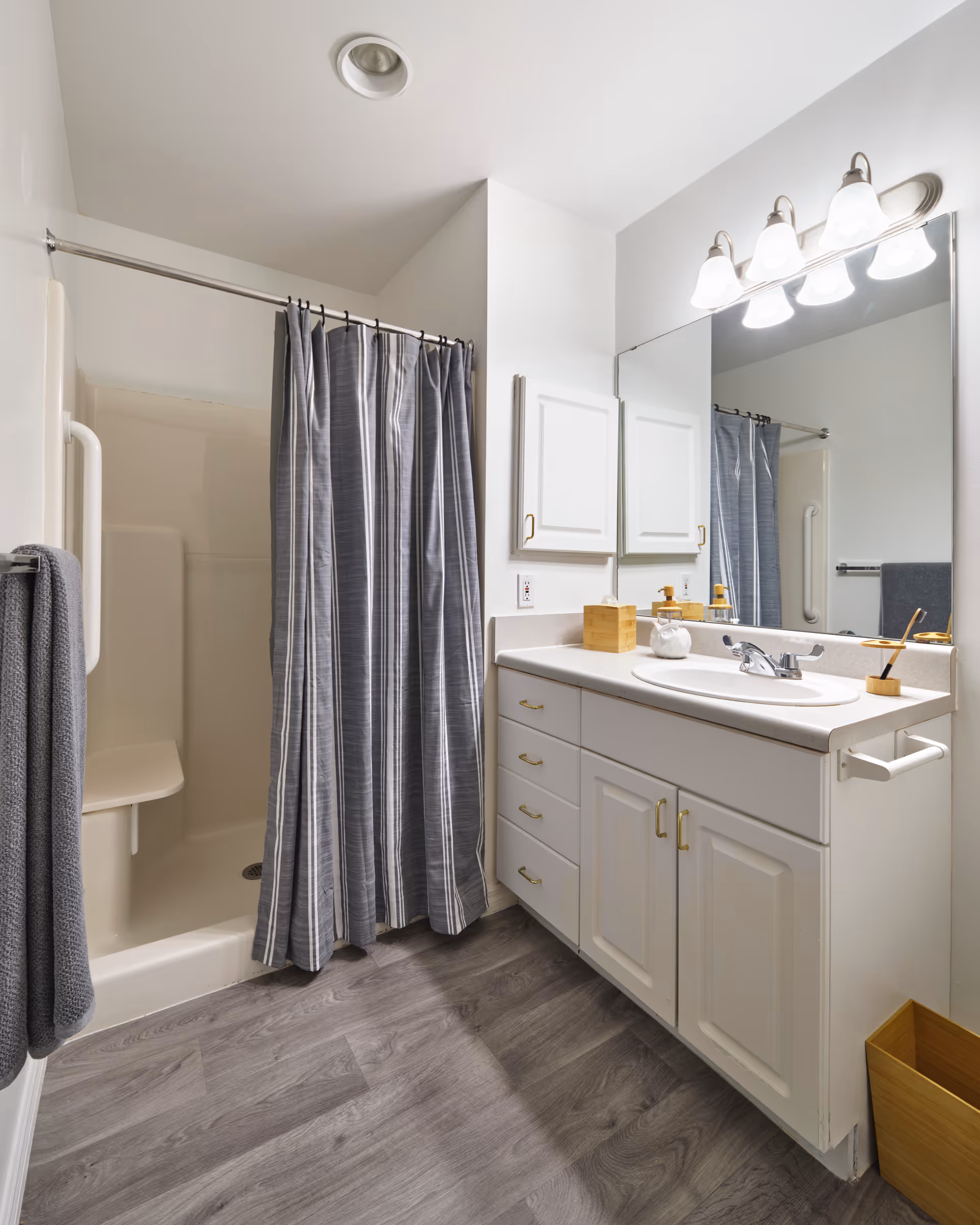 A clean and modern bathroom featuring a shower with a gray striped curtain, a white vanity with multiple drawers and cabinets, a large mirror above the sink, and a set of four light fixtures mounted on the wall. The floor has gray wood-like vinyl planks, and there is a gray towel hanging on a rack beside the shower.