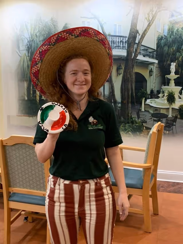 A woman with curly red hair wearing a large colorful sombrero and a dark green polo shirt with Ville Ste Marie Senior Living logo stands indoors holding a small decorative sombrero painted with the colors of the Mexican flag. Behind her are wooden chairs with blue cushions and a large wall mural depicting an outdoor courtyard with trees, a fountain, and patio furniture.