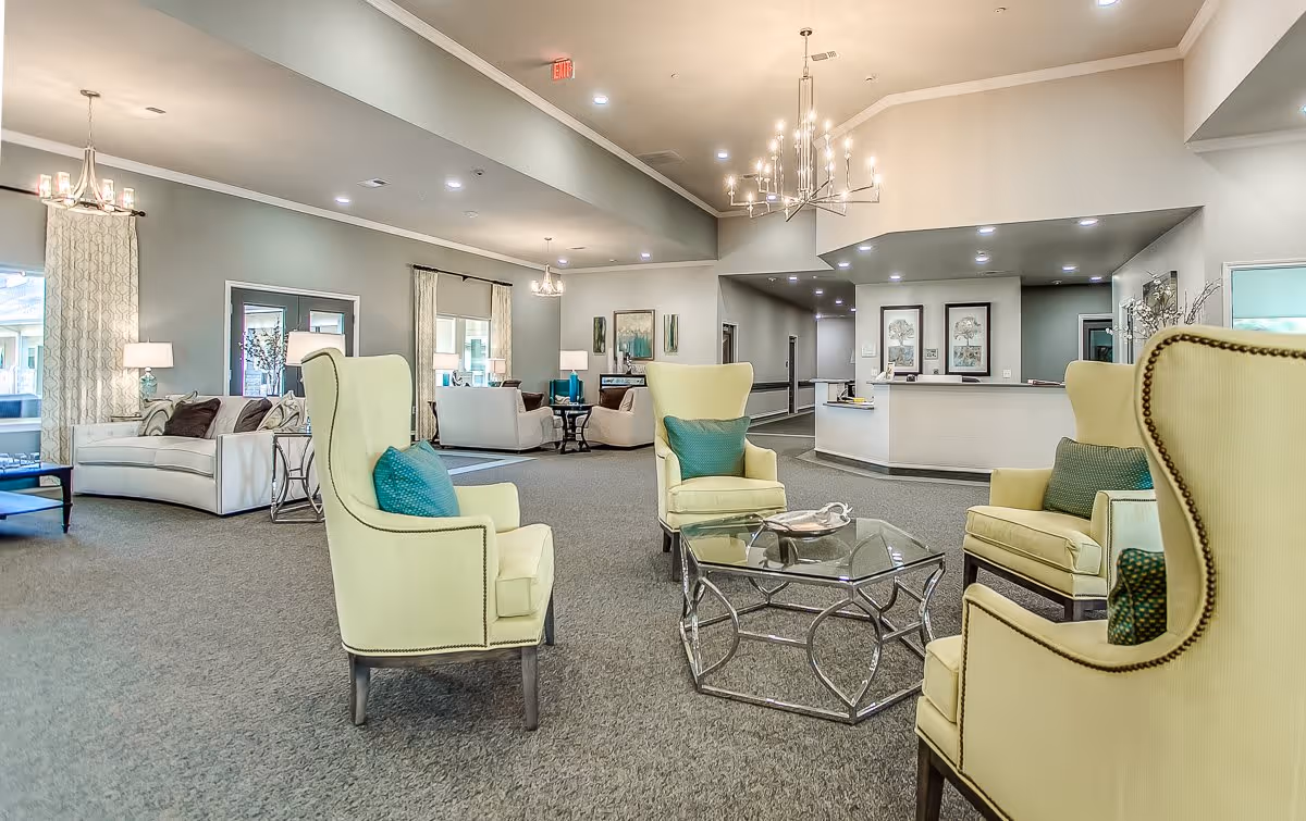 A spacious senior living facility common area with light green armchairs arranged around a glass coffee table, beige sofas, decorative pillows, chandeliers, and a reception desk in the background.