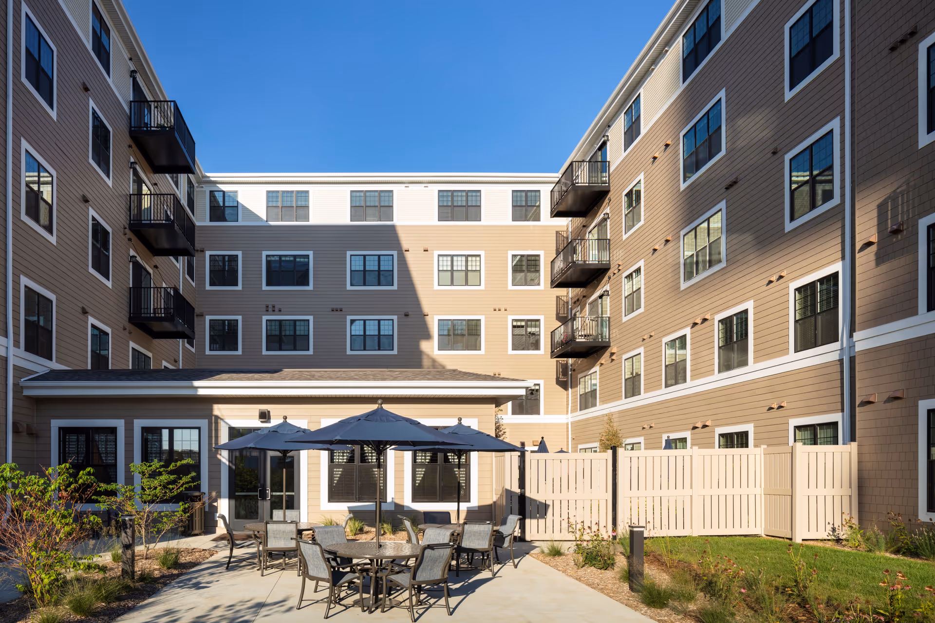 Outdoor courtyard area of a senior living facility with patio tables, chairs, and umbrellas surrounded by a multi-story building with balconies and numerous windows under a clear blue sky.