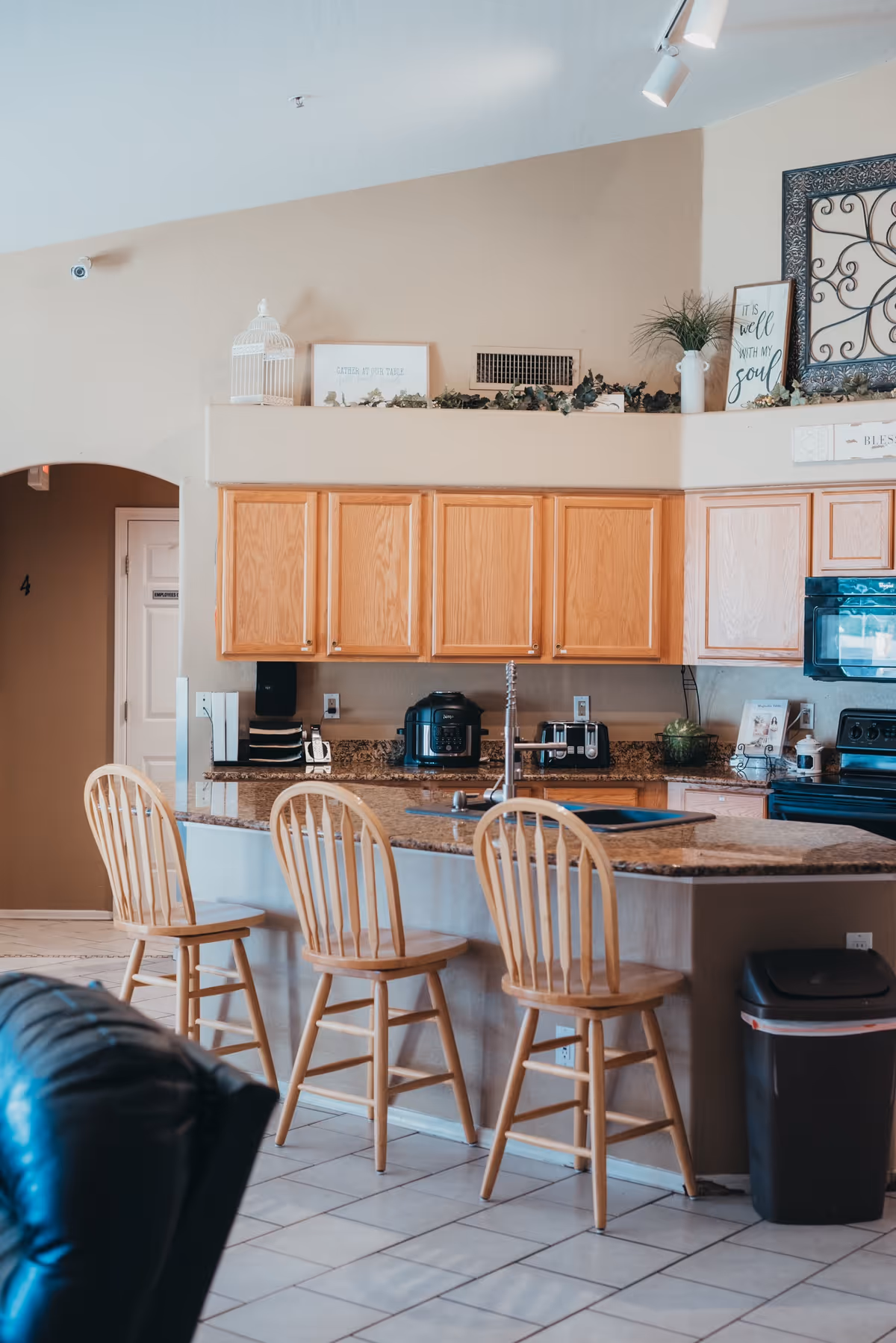 A kitchen area with a granite countertop island featuring three wooden bar stools. The kitchen has light wood cabinets, a black microwave, a black stove, and various small appliances on the counter. Decorative items and framed signs are placed above the cabinets. The floor is tiled, and part of a black leather chair is visible in the foreground.