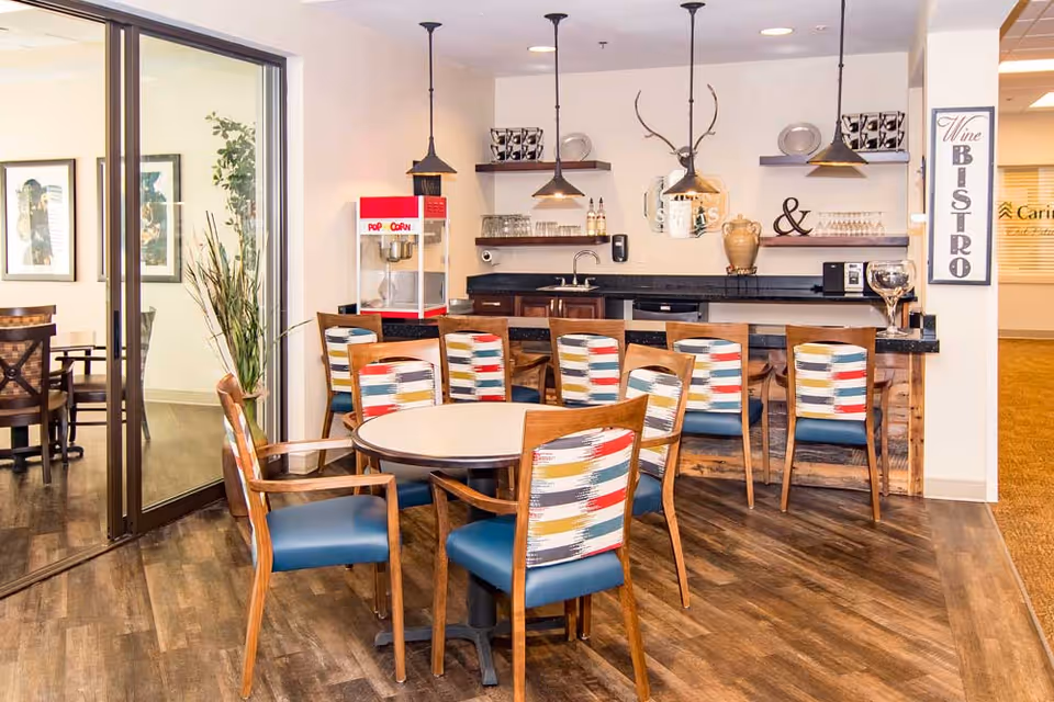A cozy dining area with a round table surrounded by six wooden chairs with colorful patterned backs and blue seats. Behind the table is a bar counter with six matching chairs. Above the counter are three pendant lights, shelves with glassware, decorative items, and a popcorn machine. A sign on the wall reads 'Wine Bistro'. The floor is wood, and there is a glass door leading to another room with additional seating and framed artwork on the walls.
